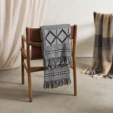 Gutte Geometric Throw in black with diamond and checked woven pattern and fringed ends, draped over a wooden chair with leather straps in a room setting.