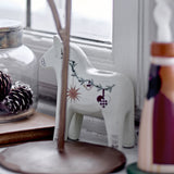 White resin Dala horse tealight holder with painted Christmas motifs and candle socket on back, shown on a windowsill with pinecones and jar.