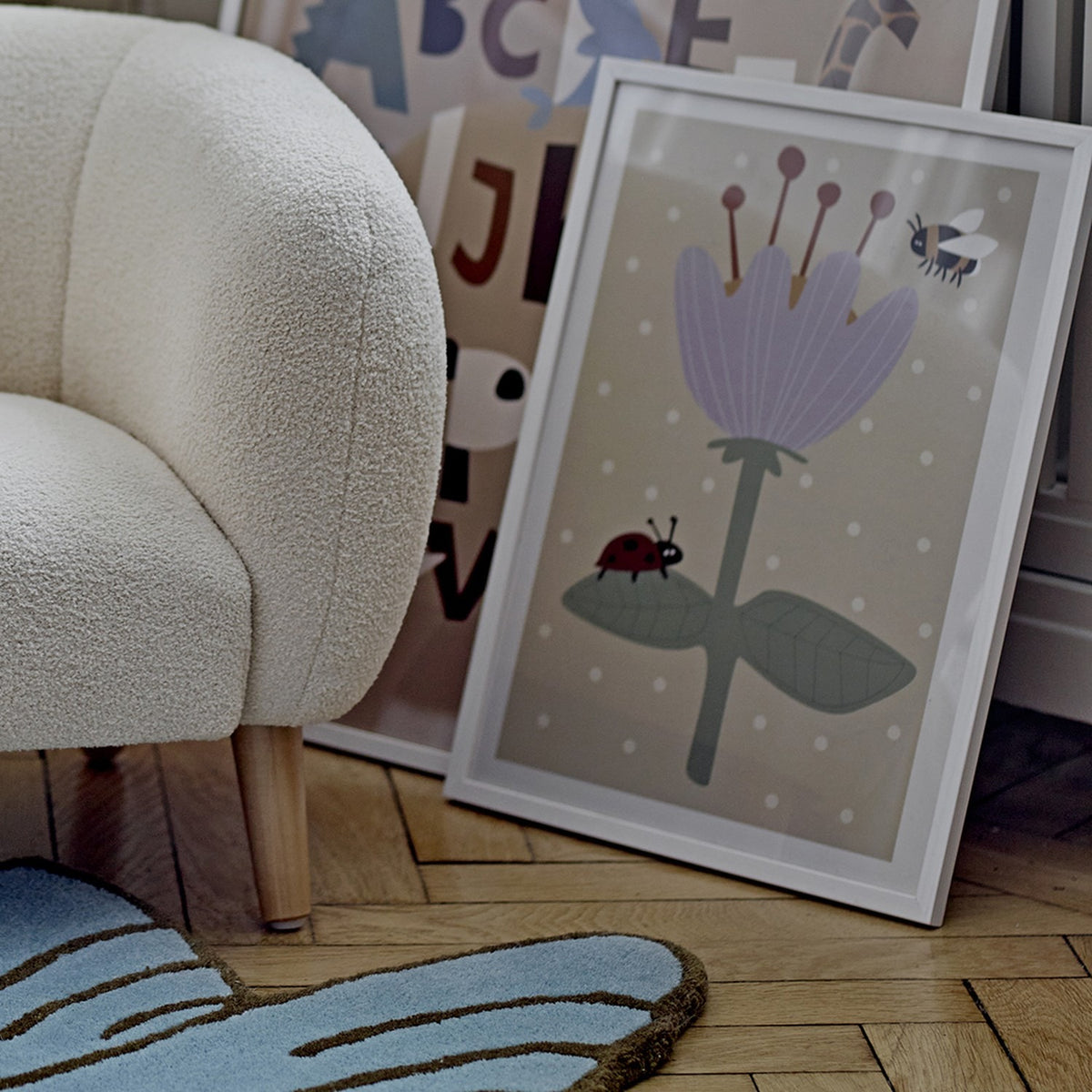 Numa Wooden Framed Print in pine, shown leaning against a wall beside a cream bouclé armchair, featuring a lilac flower, bee and ladybird on a spotted beige background.