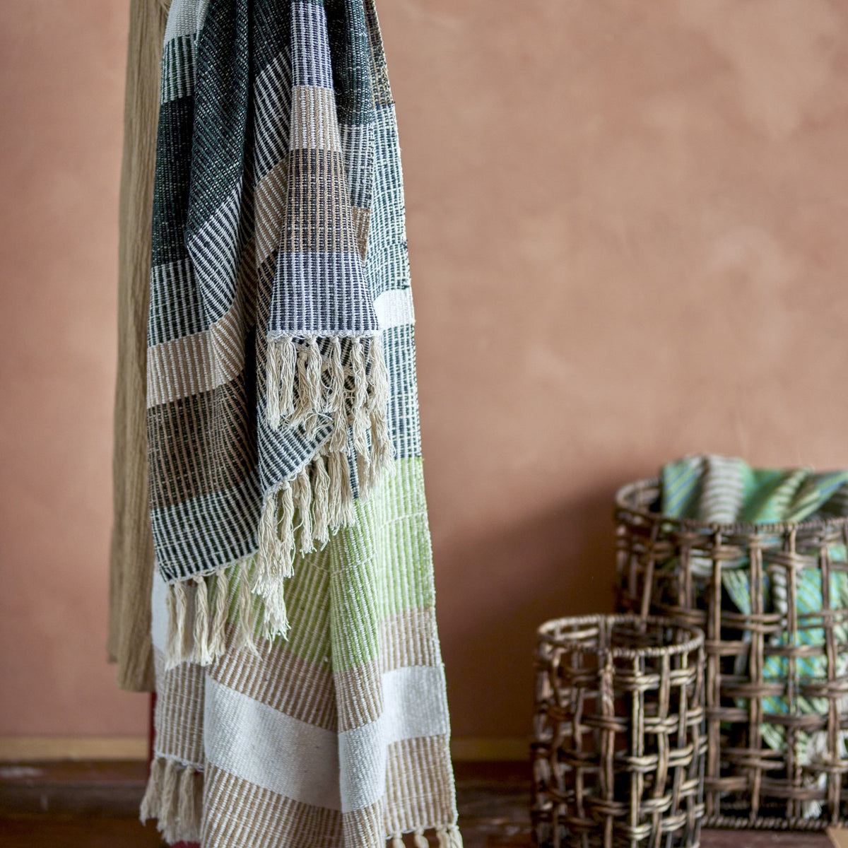 Isnel Black Striped Cotton Throw in multicoloured stripes with knotted fringe, hanging to show ribbed woven texture, styled against a terracotta wall with woven baskets nearby.