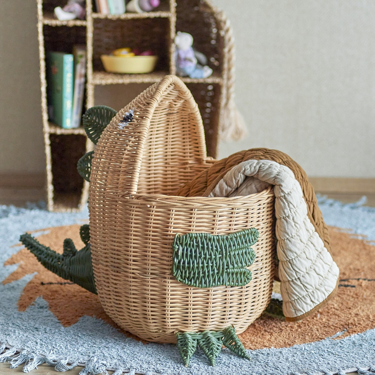 Eddi Dinosaur Rattan Basket in natural with green woven spines, arm and clawed feet, open to show a quilted blanket inside, set on a rug near a bookshelf.