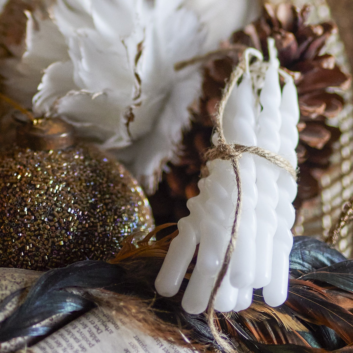 Bundle of white spiral twist candles tied with twine, resting among pine cones, feathers, and gold baubles in a decorative display.