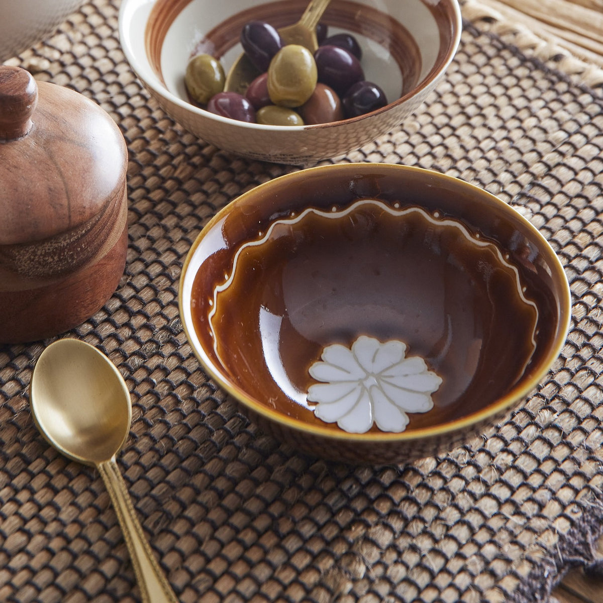 Heikki Floral stoneware bowl in brown with a white flower motif and wavy rim line, shown with a bowl of olives, gold spoon, and wooden grinder.