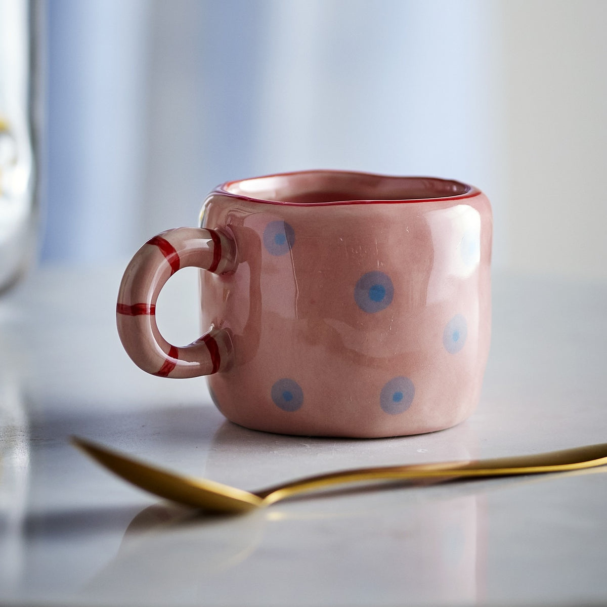 Nini Dotted Stoneware Cup in pink, small ceramic cup with hand-painted blue dots and a striped ring handle, shown on a marble surface with a gold spoon.