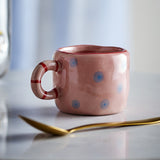 Nini Dotted Stoneware Cup in pink, small ceramic cup with hand-painted blue dots and a striped ring handle, shown on a marble surface with a gold spoon.