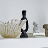 Bijan black resin candle holder with open oval loop and flared neck, shown beside a glass bowl, sculptural bust, and stone object on a white shelf.