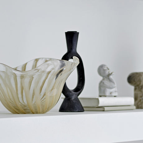 Bijan black resin candle holder with open oval loop and flared neck, shown beside a glass bowl, sculptural bust, and stone object on a white shelf.