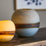 Melike Striped Glass Vase 18.5cm in Blue, a round frosted glass vase with a chalky stone-like finish and a dark uneven band at its middle, shown on a wooden surface beside a glowing candle holder.