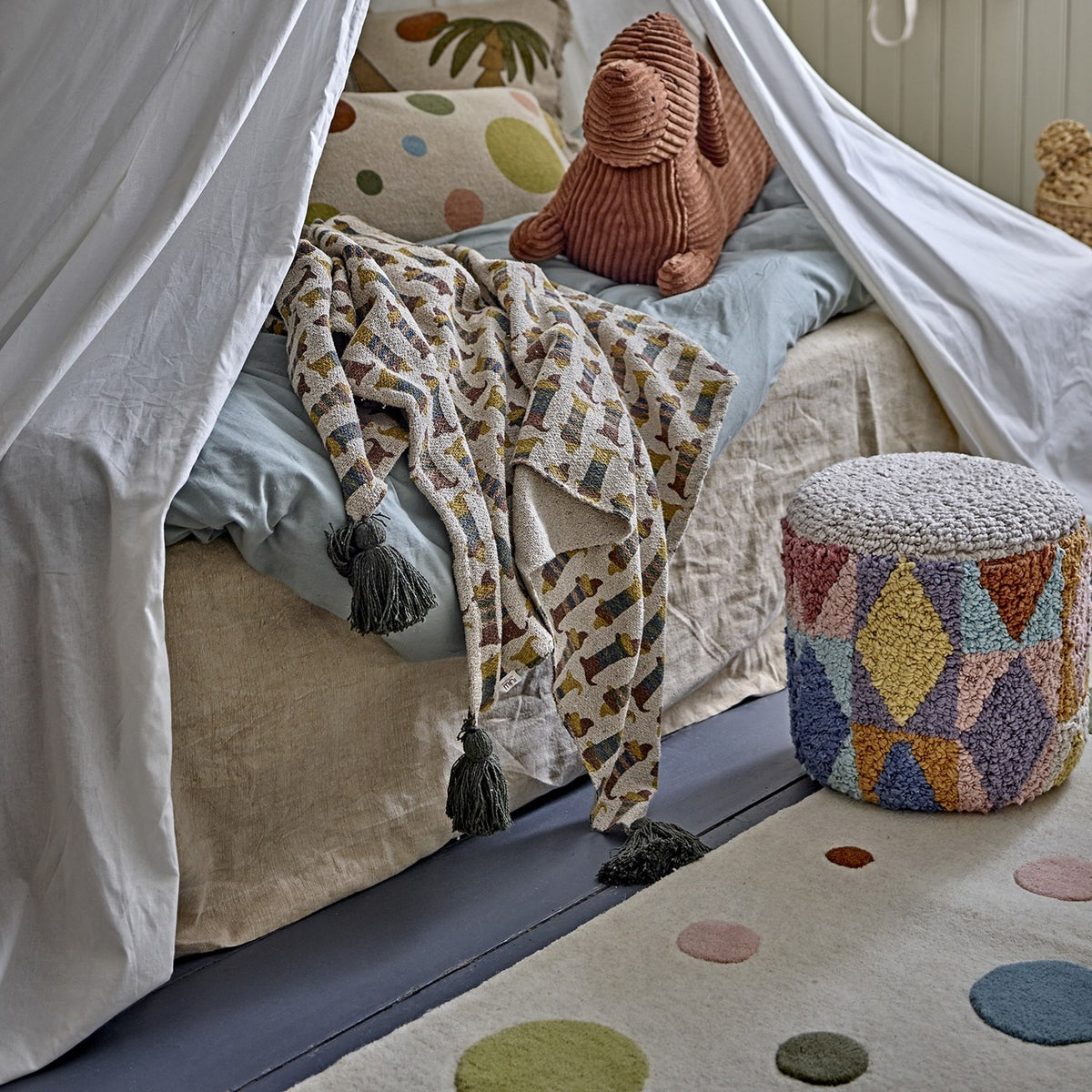 Charlie Cotton Throw in multicoloured sausage dog print with tasselled ends, draped over a bed beside a corduroy rabbit toy in a canopy-draped children's room.