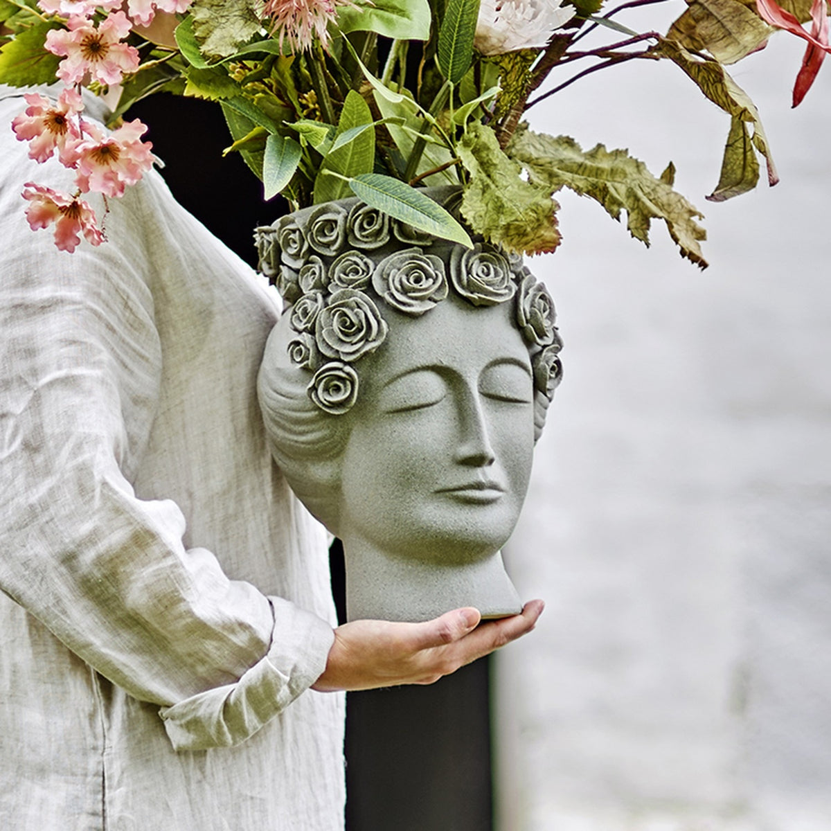 Akira Stoneware Vase 30cm in grey, sculpted as a serene woman's head with a rose wreath rim, held in hand and filled with greenery and pink blooms.