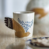 Hezha Stoneware Cup 260ml in cream, hand-painted with a bird in flight whose fanned tail forms the handle, with blue floral sprigs on a cream ground.