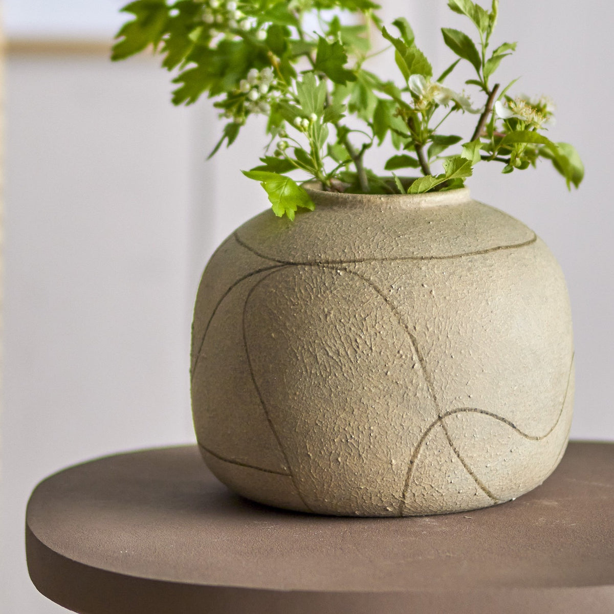 Niza Terracotta Vase 16.5cm in natural, a round, textured vase with a narrow raised-rim neck and etched curved line pattern, shown holding green foliage on a dark side table.