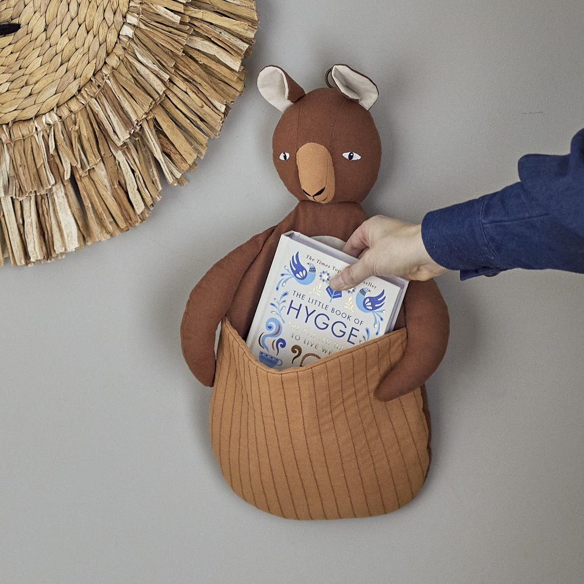 Toto Linen Wall Basket in brown, a kangaroo-shaped fabric storage holder with quilted front pouch, shown mounted on a wall holding a book as a hand reaches inside.