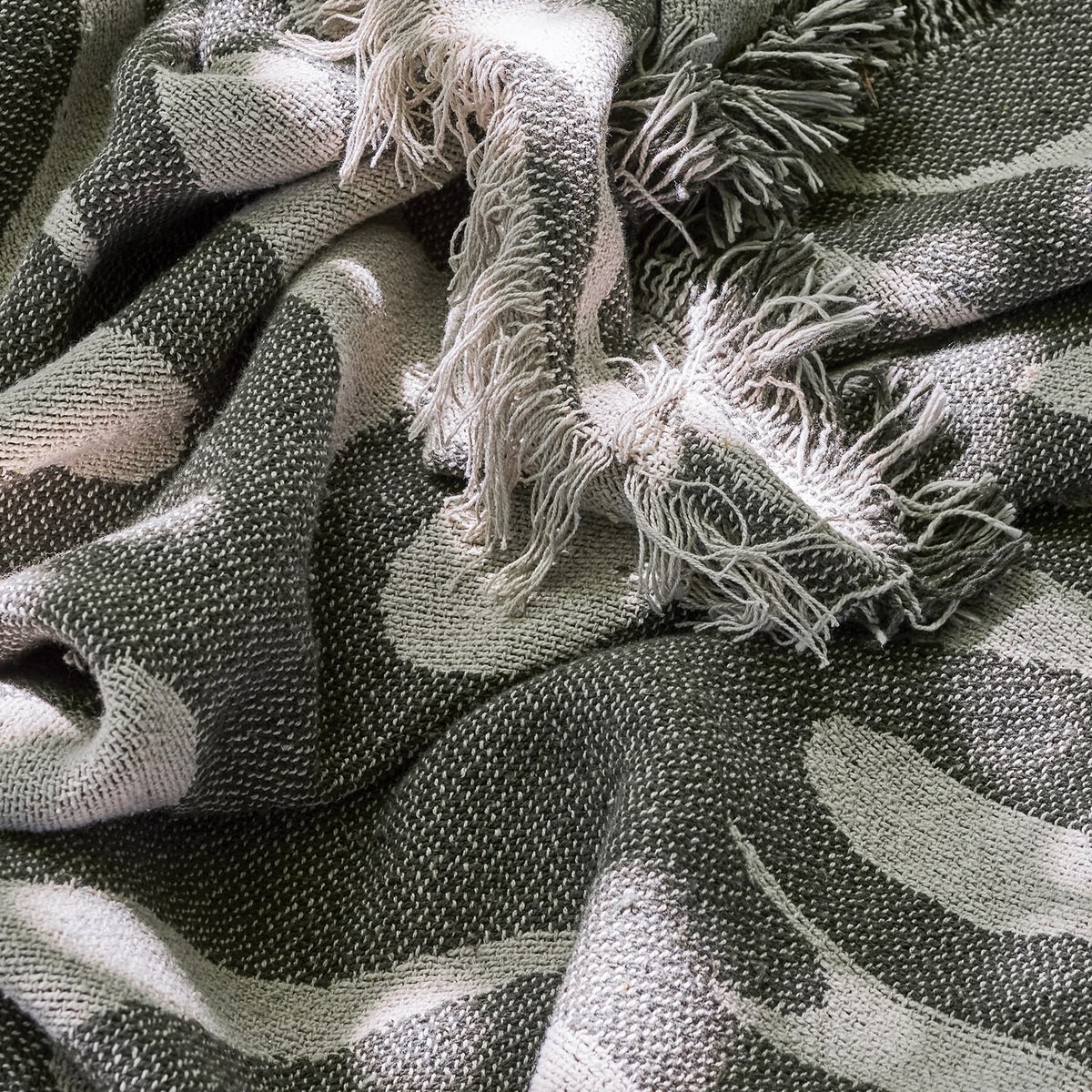 Alk Cotton Throw in green, woven cotton blend fabric with a rounded leaf-like pattern, shown draped with loose fringed edges visible.