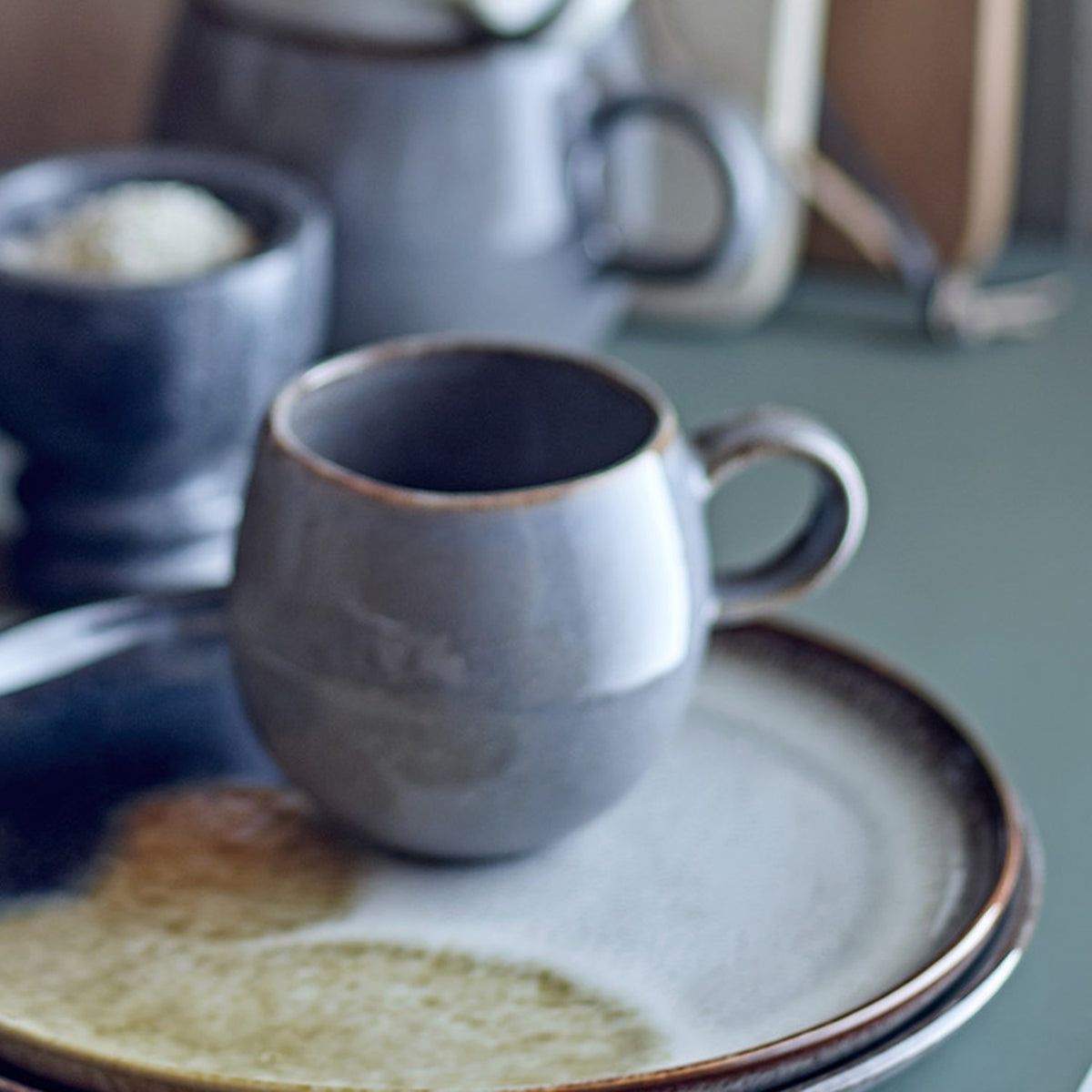 Sandrine Stoneware Cup 275ml in grey with a round, ball-shaped body, loop handle, and speckled reactive glaze darkening towards the rim, shown on a matching stoneware plate.