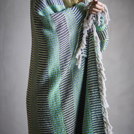 Isnel Green Striped Throw draped over a person's arm, showing fine multicoloured stripes, ribbed woven texture, and a fringed edge against a plain grey background.