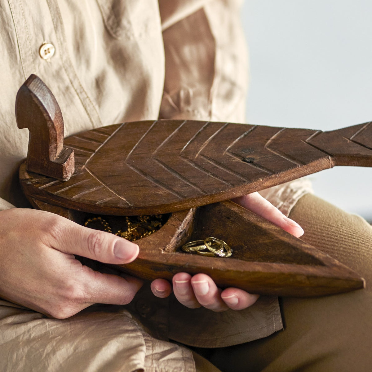 Hamida fish-shaped wooden storage box in brown with carved chevron scale lid, shown open in hand to reveal rings and a chain necklace inside.