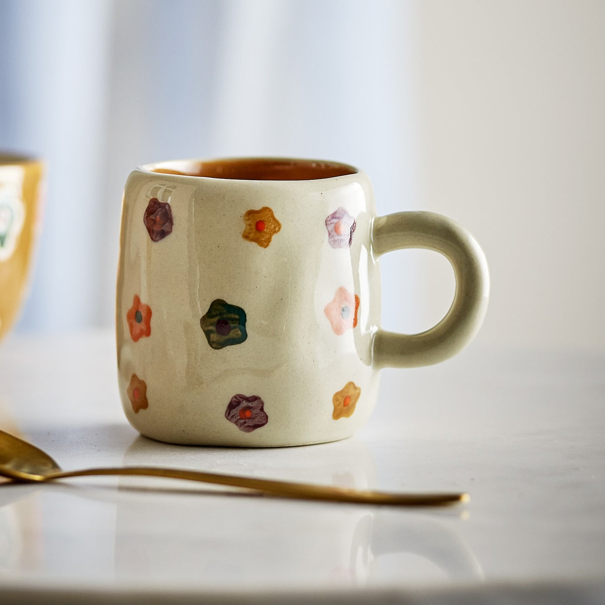 Addy Stoneware Cup 130ml in cream, a small rounded ceramic cup with a loop handle, hand-painted with scattered multicoloured flowers, shown on a marble surface with a gold spoon.
