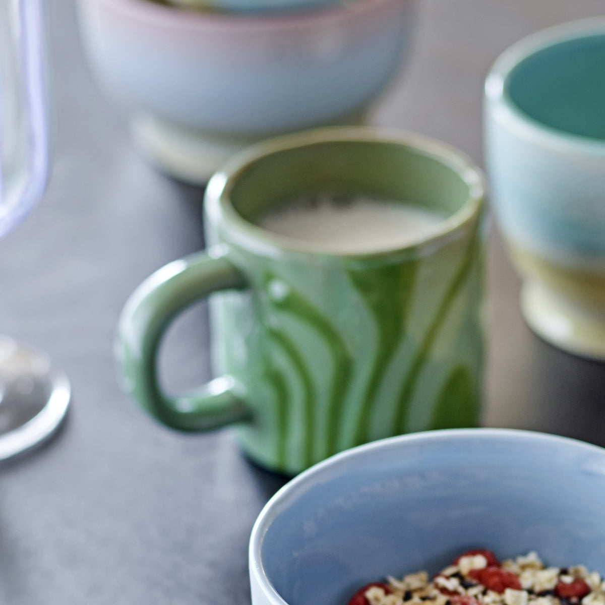 Ninka Stoneware Cup in green with bold striped glaze and rounded loop handle, filled with milk, shown among blurred ceramic bowls on a grey surface.