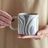Hands holding the Ninka stoneware cup in grey, showing its rounded loop handle and flowing curved bands against a neutral backdrop.