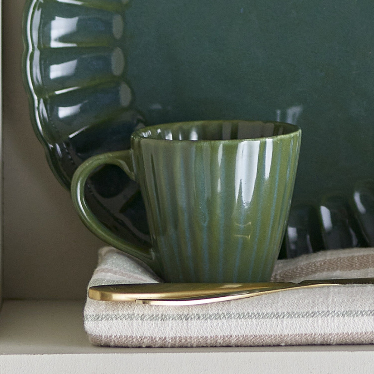 Latina Stoneware Cup 220ml in green with fluted, ribbed sides and an open loop handle, shown beside a dark green scalloped plate and striped linen napkin.
