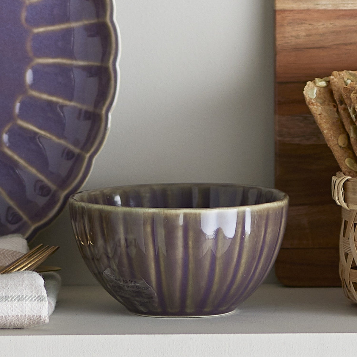 Latina Stoneware Bowl 9cm in grey with fluted ribbed sides, shown on a shelf beside a purple scalloped plate and crackers.