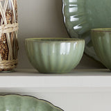 Latina Stoneware Bowl 12cm in green, round with fluted sides and a wavy rim, shown on a shelf among matching green ceramic plates and bowls.