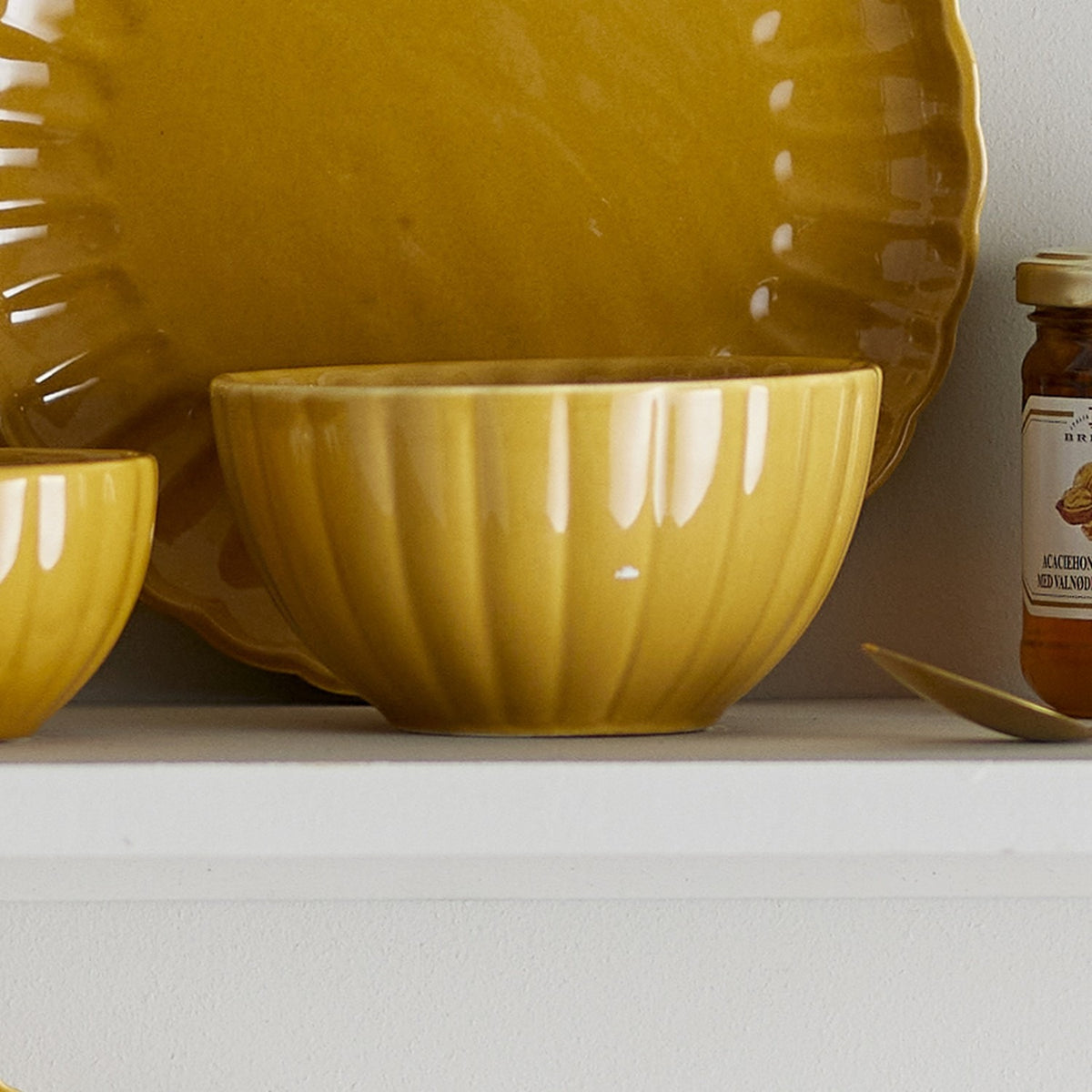 Latina Stoneware Bowl 12cm in yellow, fluted ceramic sides with a glossy glaze, shown on a shelf beside matching yellow plates and a small jar.