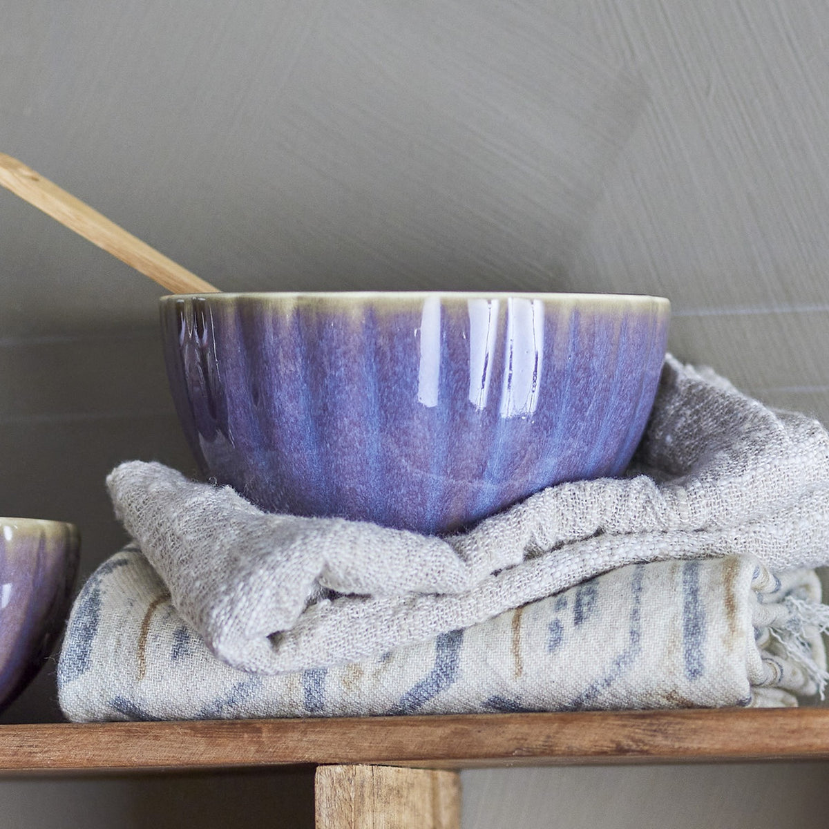 Latina Stoneware Bowl 12cm in purple with fluted sides and glossy glaze, shown with a wooden spoon on folded linen cloths against a grey background.