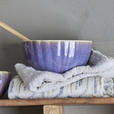 Latina Stoneware Bowl 12cm in purple with fluted sides and glossy glaze, shown with a wooden spoon on folded linen cloths against a grey background.