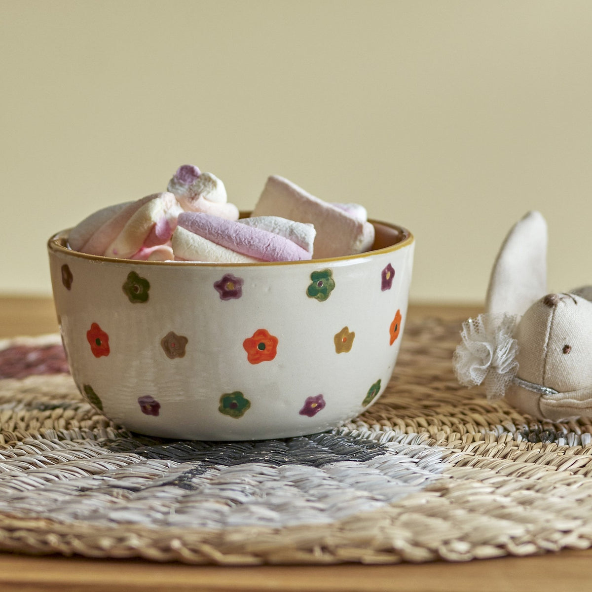 Addy Stoneware Bowl 12cm in cream, filled with marshmallows, featuring hand-decorated multicoloured flowers and a contrasting rim, shown on a woven mat with a plush toy nearby.