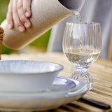 Taurin Round Wine Glass in clear ribbed glass with a short round foot, shown being filled with white wine at an outdoor table setting with plates.