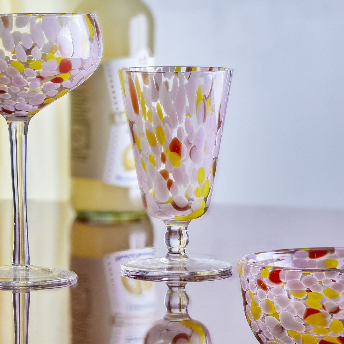 Lilya Speckled Wine Glass in Pink, a stemmed glass with flared bowl, rounded knop and flat foot, shown with a matching coupe and bowl on a reflective surface.