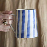 Hand with a gold ring holding the Begonia Striped Stoneware Mug 440ml in blue, showing hand-painted vertical stripes on a ceramic mug against a beige backdrop.
