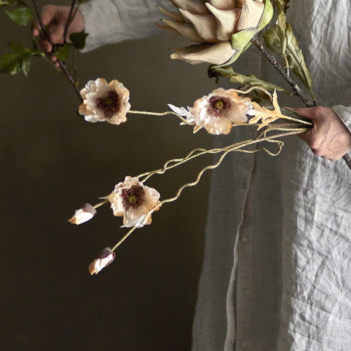 Poppy Artificial Stem in beige held alongside dried botanicals, showing wavy branching stems with open ruffled poppy flowers, closed buds, and small leaves.