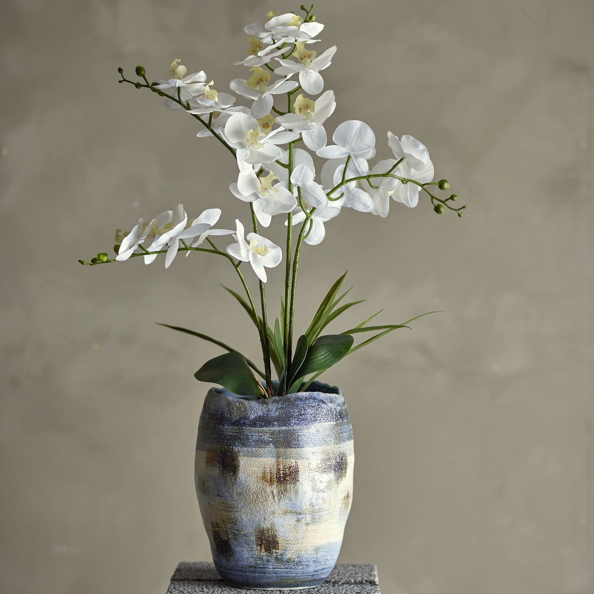 Artificial white orchid plant with arching stems and broad leaves in a blue-grey patterned pot, set against a neutral background.