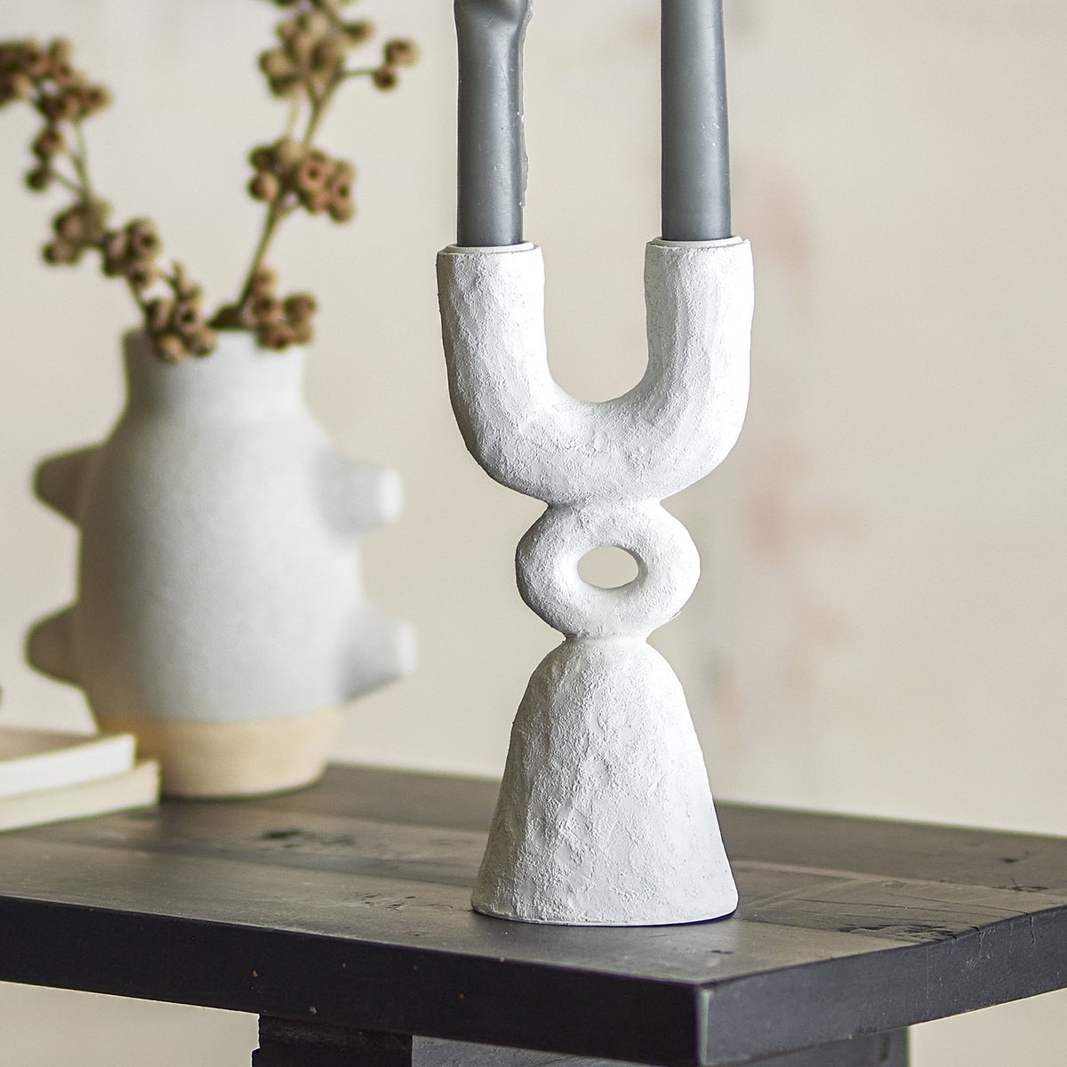 Syra Resin Candle Holder 22cm in white, a sculptural two-arm design with an open ring detail and flared base, holding grey candles on a dark shelf beside a textured vase.