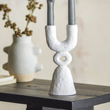 Syra Resin Candle Holder 22cm in white, a sculptural two-arm design with an open ring detail and flared base, holding grey candles on a dark shelf beside a textured vase.