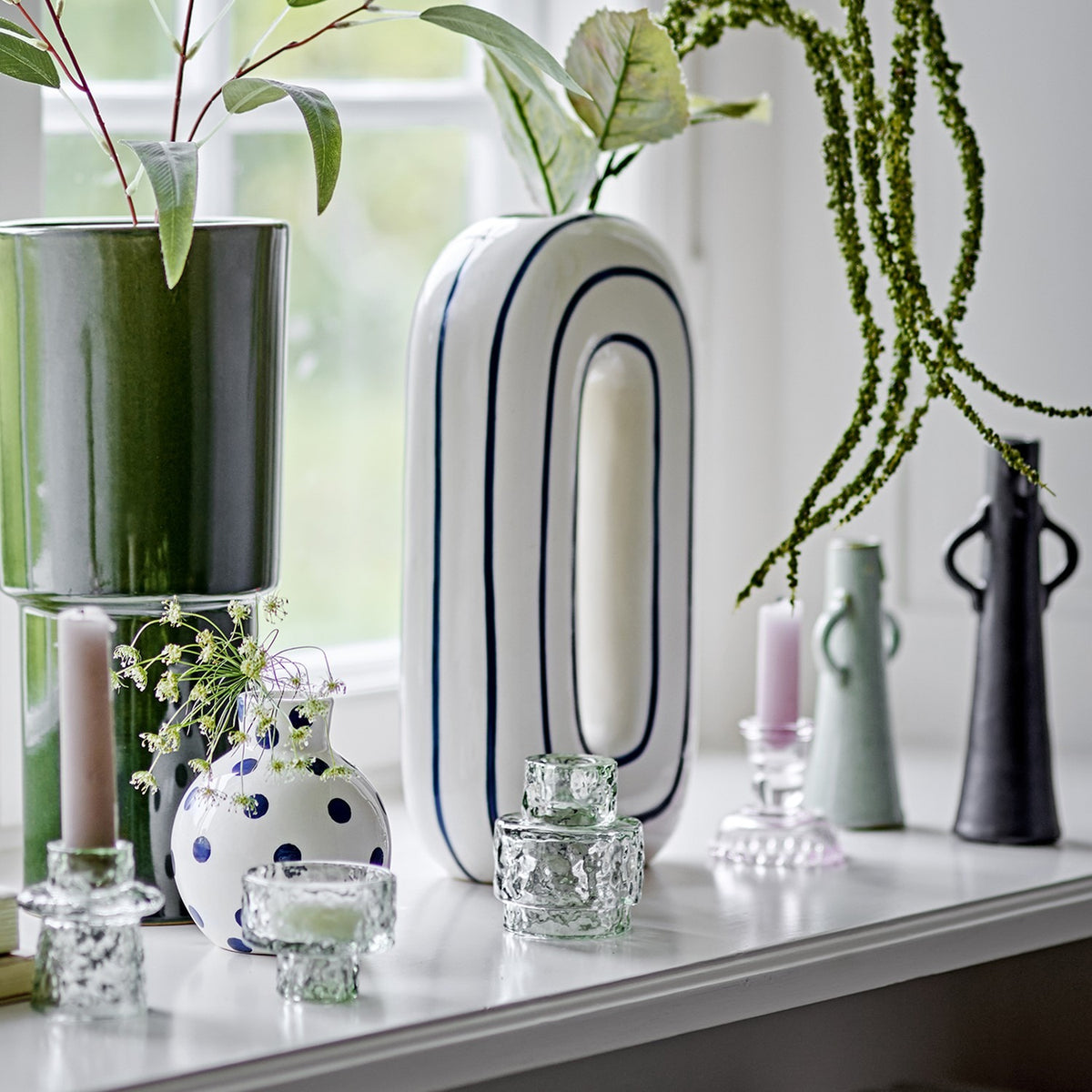 Mae Striped Ceramic Vase 32cm in White, a tall oval loop-shaped vase with blue outline stripes, shown on a shelf among candle holders and greenery near a window.