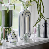 Mae Striped Ceramic Vase 32cm in White, a tall oval loop-shaped vase with blue outline stripes, shown on a shelf among candle holders and greenery near a window.