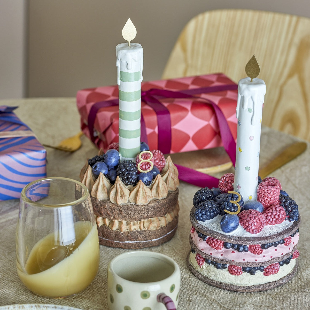 Hooray resin birthday decoration cakes with berry toppings and gold-flame candles, shown on a table with gift boxes, a mug, and a glass of juice.