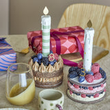 Hooray resin birthday decoration cakes with berry toppings and gold-flame candles, shown on a table with gift boxes, a mug, and a glass of juice.