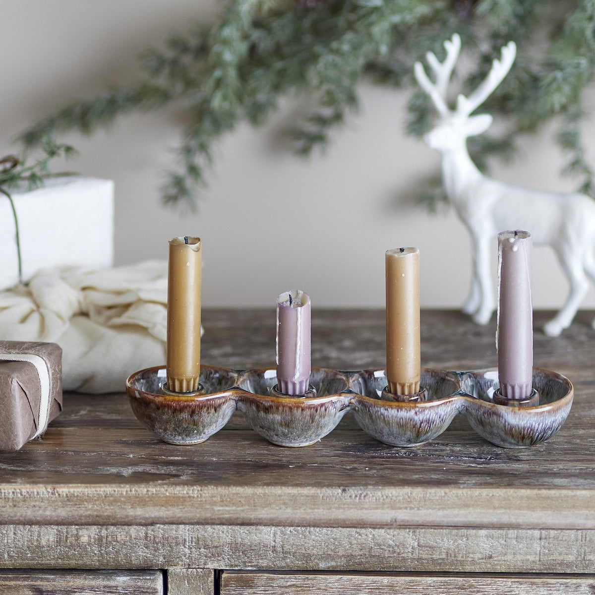 Kalia Stoneware Advent Candle Holder in brown reactive glaze with four joined round cups holding tan and mauve dinner candles, shown on a wooden sideboard with festive decor.