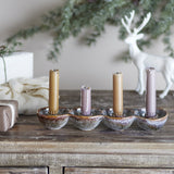Kalia Stoneware Advent Candle Holder in brown reactive glaze with four joined round cups holding tan and mauve dinner candles, shown on a wooden sideboard with festive decor.