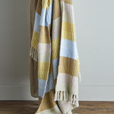 Isnel Yellow Striped Throw draped over a chair, showing a woven multicoloured striped pattern in mustard, blue, and cream with knotted tassel fringe.