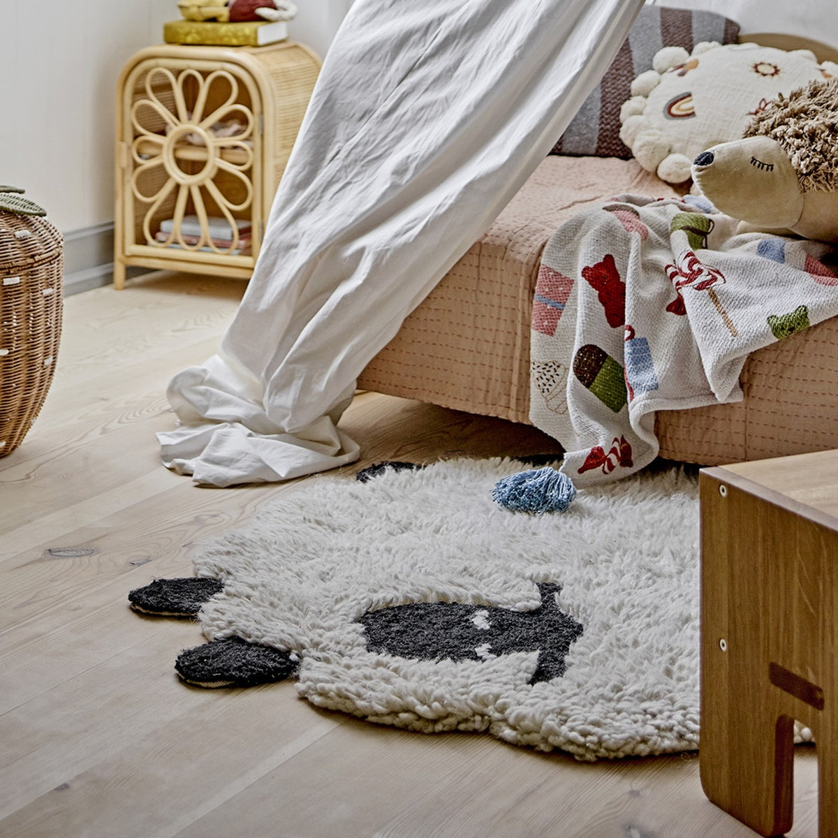 Dolly Sheep Wool Rug in cream, 84x84cm, shaped like a sleeping sheep with curly pile, dark face and feet, shown on a nursery floor beside a bed and rattan furniture.