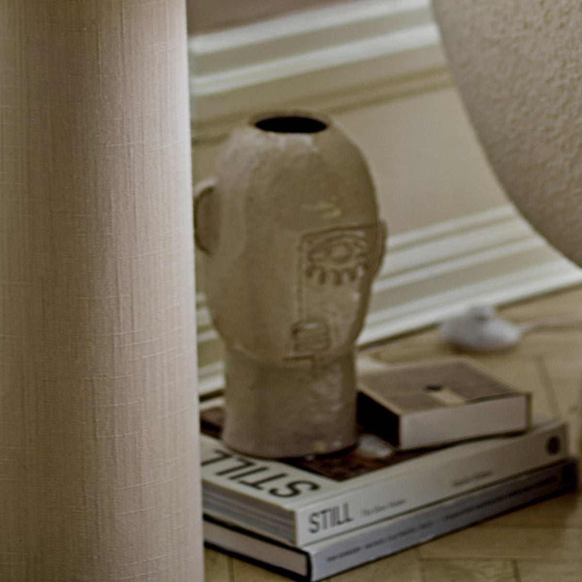 Lizzie Stoneware Vase 25.5cm in beige, shaped like a head with ears and a raised-line face, displayed on books beside a textured lamp base and curtain.