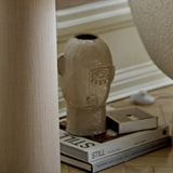 Lizzie Stoneware Vase 25.5cm in beige, shaped like a head with ears and a raised-line face, displayed on books beside a textured lamp base and curtain.