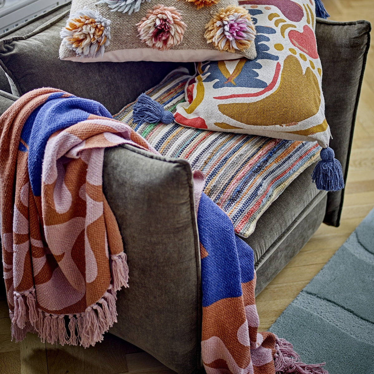 Atle Leaf Cotton Throw in brown with pink fringed edges, draped over a grey armchair alongside patterned cushions, showing its botanical print and blue accent tones.