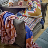 Atle Leaf Cotton Throw in brown with pink fringed edges, draped over a grey armchair alongside patterned cushions, showing its botanical print and blue accent tones.